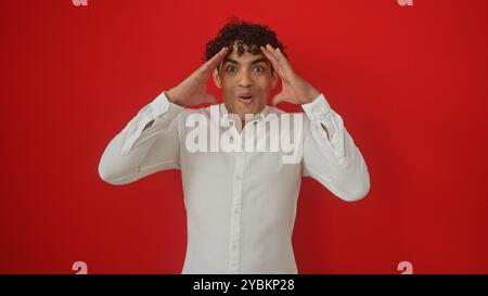 Surpris jeune homme hispanique en chemise blanche sur fond rouge solide, montrant l'excitation Banque D'Images