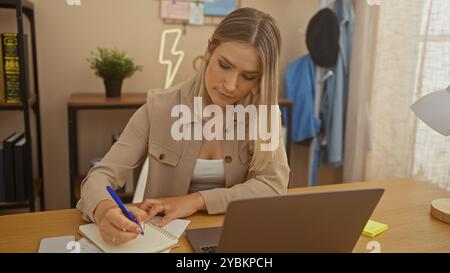 Une jeune femme blonde travaille avec diligence à son bureau dans un bureau à domicile moderne, entourée de livres et de décors, prenant des notes et utilisant un ordinateur portable dans un organi Banque D'Images