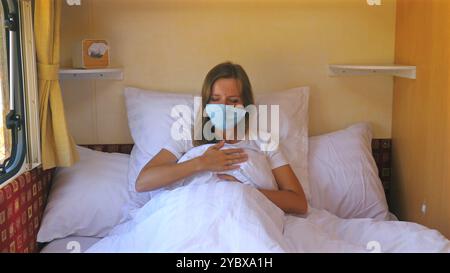 Femme portant un masque facial, assise sur le lit et toussant, à l'intérieur d'une maison confortable Banque D'Images