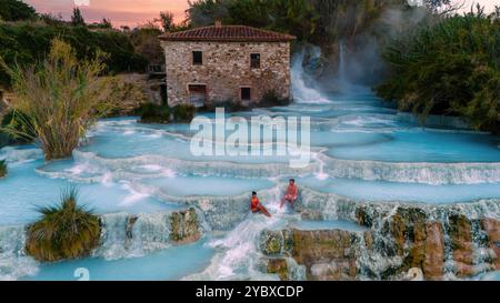 Les visiteurs apprécient les eaux apaisantes des bains thermaux Saturnia en Toscane, entourés par une nature luxuriante et une vapeur chaude s'élevant sous le SK coloré du soir Banque D'Images