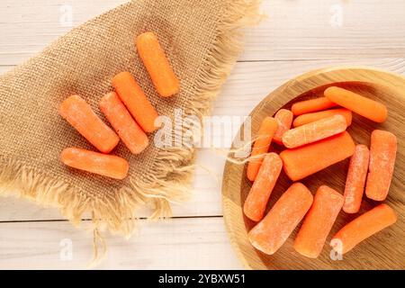 Petites carottes pelées avec des ustensiles en bois et serviette en jute sur une table en bois, macro, vue de dessus. Banque D'Images