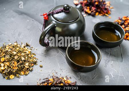 Thé sec de différentes variétés et une théière avec des tasses sur un fond gris. Cérémonie du thé, thé avec divers additifs, mélange. Photo de haute qualité Banque D'Images