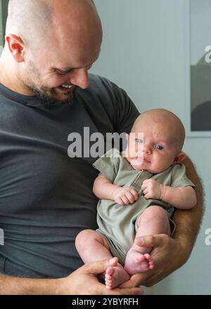 Père tient amoureusement son fils nouveau-né dans les bras, le regardant affectueusement. Bébé repose paisiblement dans les bras de son père. Atmosphère chaleur et paix. Banque D'Images