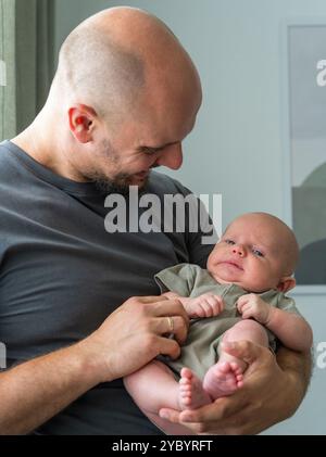 Père tient amoureusement son fils nouveau-né dans les bras, le regardant affectueusement. Bébé repose paisiblement dans les bras de son père. Atmosphère chaleur et paix. Banque D'Images