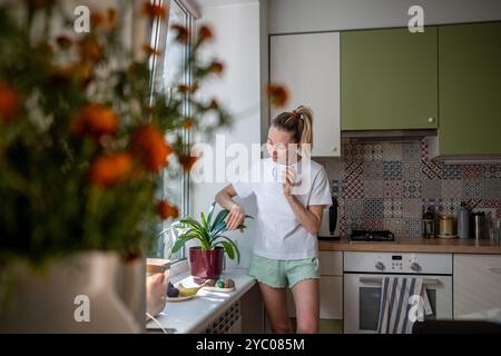 Sereine femme au foyer arrosant les plantes d'intérieur, vérifiant la santé des fleurs, dépoussiérant les feuilles dans la cuisine seule Banque D'Images