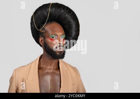 Modèle gay afro-américain confiant avec maquillage sur le visage portant une perruque énorme sur fond gris. L'homme noir porte un blazer en studio. Banque D'Images