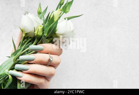 Une main avec de longs ongles élégants tient un bouquet de fleurs blanches fraîches. Les verts et les blancs doux contrastent magnifiquement avec un dos texturé léger Banque D'Images