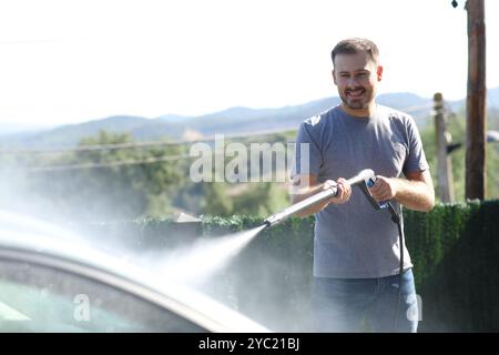 Homme heureux lavant voiture avec tuyau à haute pression dans le jardin à la maison Banque D'Images