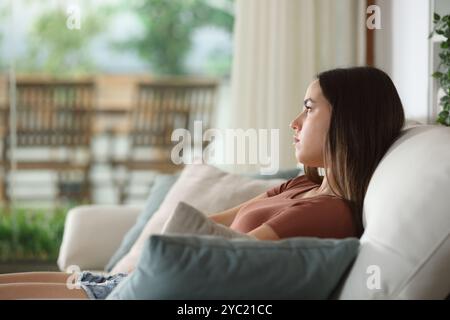 Femme sérieuse regardant loin par une fenêtre assise sur un canapé à la maison Banque D'Images