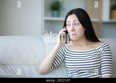 Femme asiatique perplexe parlant au téléphone assis sur un canapé à la maison Banque D'Images