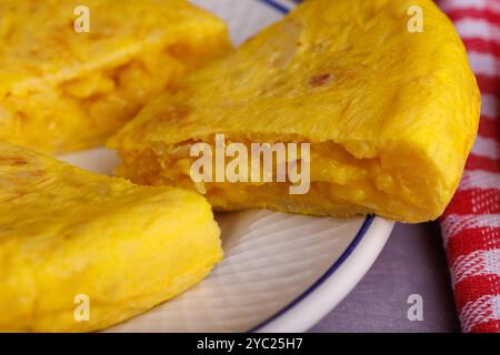 Gros plan d'une tranche d'omelette de pomme de terre espagnole à côté d'une serviette à carreaux rouge, du pain frais et des œufs sur une table en béton Banque D'Images