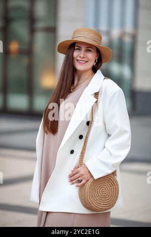 Joyeux sourire élégant femme taille plus debout à l'extérieur dans la ville urbaine, portant une robe beige, blazer blanc, chapeau à large bord, sac à main tissé. Modèle avec Banque D'Images