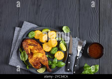 la moitié de poulet rôti avec des pièces d'épis de maïs et des feuilles de basilic frais sur une assiette sur une table en bois noir avec des couverts et une trempette au chili doux, horizontal vi Banque D'Images