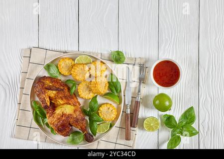 la moitié du poulet bbq avec des pièces d'épis de maïs et des feuilles de basilic frais sur une assiette sur une table en bois blanc avec couverts, citron vert et sauce chili doux, horizontal vi Banque D'Images