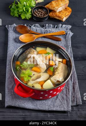 Soupe de brochet clair avec légumes, persil et épices dans un pot rouge sur une table en bois noir avec des cuillères en bois et du pain grillé, vue verticale Banque D'Images