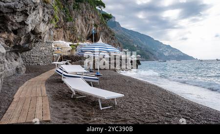 Les visiteurs se détendent le long de la plage parsemée de galets de la côte amalfitaine, en profitant d'une vue magnifique, de parasols rayés et de vagues tranquilles lançant sur le shor Banque D'Images