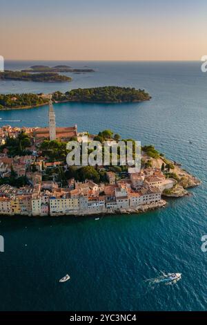 Les eaux tranquilles de Rovinj scintillent au coucher du soleil, projetant une lueur chaude sur les bâtiments historiques et la végétation luxuriante. Les bateaux glissent doucement, enhan Banque D'Images