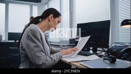 Belle femme adulte spécialiste financier travaillant avec des papiers, recherchant et analysant des données, des graphiques et des graphiques de métriques financières changeant sur le moniteur d'ordinateur. Travailler dans un bureau d'affaires moderne. Banque D'Images