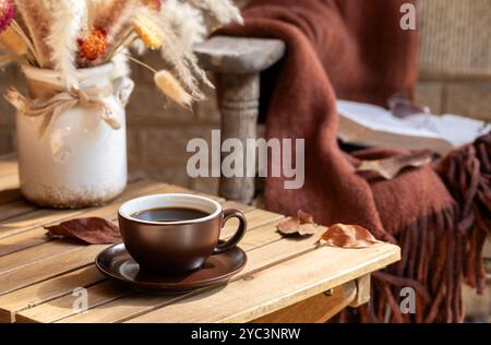 Tasse de café avec vase de fleurs d'automne séchées et de feuilles sur table en bois et vieille chaise avec couverture en arrière-plan Banque D'Images