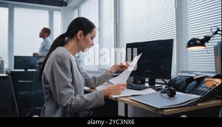 Belle femme adulte spécialiste financier travaillant avec des papiers, recherchant et analysant des données, des graphiques et des graphiques de métriques financières changeant sur le moniteur d'ordinateur. Travailler dans un bureau d'affaires moderne. Banque D'Images