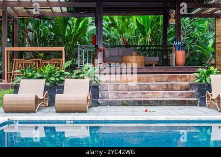 Une piscine extérieure sereine avec une piscine bleu clair, des chaises longues élégantes et une terrasse en bois entourée de plantes tropicales luxuriantes. Le réglage comprend Banque D'Images