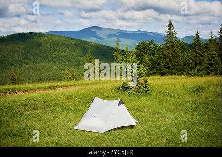 Tente touristique blanche au soleil, au sommet d'une colline herbeuse, entourée de jeunes pins. Vue panoramique sur les montagnes luxuriantes et vallonnées sous un ciel bleu vif avec des nuages dispersés, créant un endroit de camping idyllique. Banque D'Images