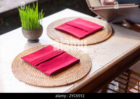 Une table de salle à manger magnifiquement sertie avec deux sets de table ronds tissés, chacun avec une serviette rouge soigneusement pliée. Un petit pot d'herbe verte ajoute une touche de Banque D'Images