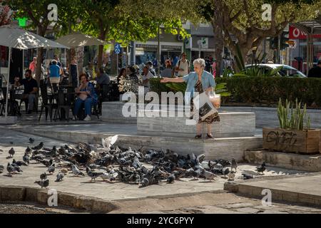 Femme âgée nourrissant des pigeons sur une place publique en Grèce, entourée de passants et de cafés. Scène de vie urbaine chaleureuse. Banque D'Images