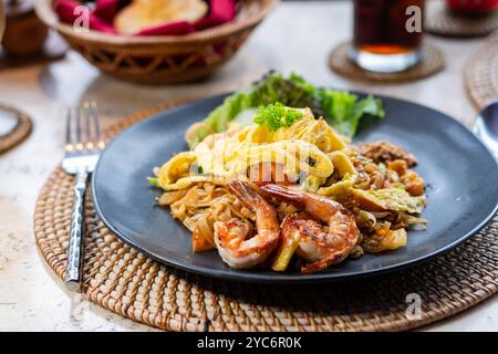 Une assiette de délicieuses nouilles sautées garnies de crevettes et d'une omelette, garnie de laitue et de légumes. Le plat est servi sur une assiette noire, Banque D'Images