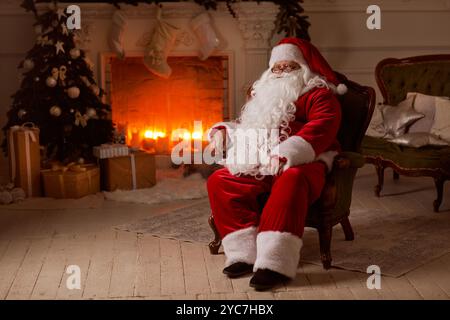 Le Père Noël est assis dans une chaise confortable près de la cheminée avec un arbre de Noël, entouré de décorations de Noël et de cadeaux. Banque D'Images