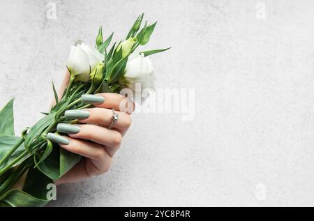 Une main avec de longs ongles élégants tient un bouquet de fleurs blanches fraîches. Les verts et les blancs doux contrastent magnifiquement avec un dos texturé léger Banque D'Images