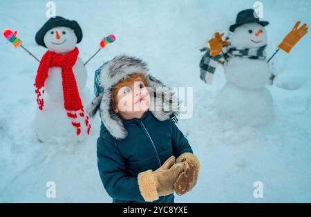 Bonhomme de neige et enfant drôle l'ami est debout dans un chapeau d'hiver et un foulard avec le nez rouge. Joyeux Noël et Bonne Année. Noël hiver enfants. Banque D'Images