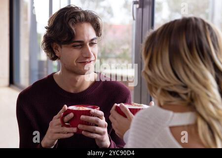 Période de Noël, couple appréciant le chocolat chaud ensemble à la maison, partageant des sourires chaleureux et la conversation Banque D'Images