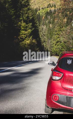 Voiture rouge avec symbole de trident ukrainien sur la route courbe dans les montagnes le jour ensoleillé. Voyage d'automne en voiture en Ukraine. Voiture à hayon sur autoroute de montagne Banque D'Images