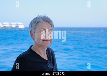 Une femme âgée aux cheveux gris courts se tient près de la mer bleue calme par une journée brillante, souriant doucement. Le fond serein de l'océan et son expressi paisible Banque D'Images