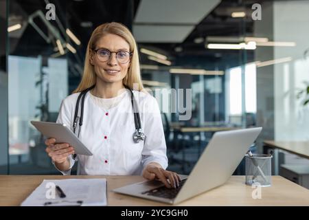 Médecin féminin mature en cabinet utilisant un ordinateur portable et une tablette pour la télémédecine. Le milieu professionnel de la santé met en avant l'expertise en technologie médicale. S Banque D'Images