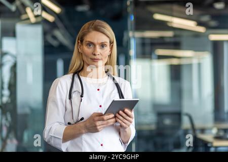 Médecin féminin mature debout en toute confiance avec tablette dans le bureau moderne. Stéthoscope autour du cou signifie le professionnalisme médical, l'accent sur le numérique Banque D'Images