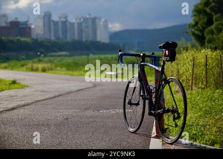 Ulsan, Corée du Sud - 9 juillet 2019 : un vélo de route repose sur le bord de la piste cyclable de la rivière Taehwa près du coucher du soleil, avec le paysage urbain au loin, blen Banque D'Images