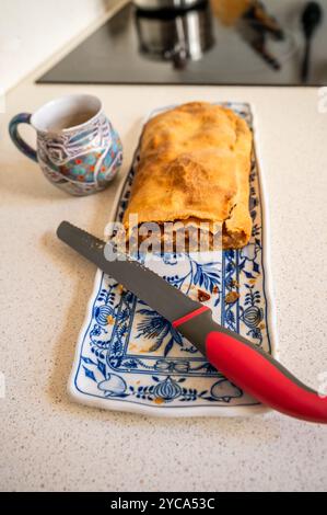 Strudel pomme sur plaque décorative bleue avec couteau rouge, tasse bleue sur plan de cuisine avec plaque de cuisson et pot en arrière-plan. Banque D'Images