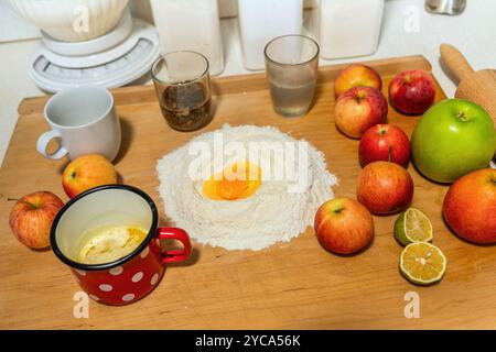 Planche de cuisine en bois avec de nombreux ingrédients (farine, oeuf, pomme, rouleau, beurre nélté dans une tasse rouge, verre, raisin, pot) pour la préparation de la cuisson du strudel de pomme. Banque D'Images