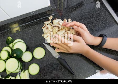 Mains de fille ramassant des ​​mushroom tranchés à côté d'un couteau et un bouquet de ​​zucchini tranchés sur un comptoir de cuisine noir à la maison Banque D'Images