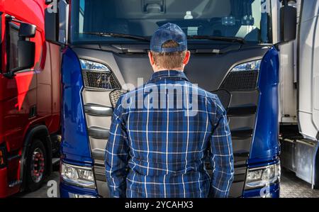 Un homme portant une casquette se tient devant un camion bleu, examinant de près ses caractéristiques tout en étant entouré d'autres grands véhicules dans une zone de transport. Banque D'Images