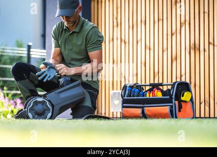 Un homme s'agenouille sur l'herbe, met des gants et se prépare avec des outils de jardinage à côté d'un sac à outils lumineux et d'une clôture en bois en arrière-plan. Banque D'Images