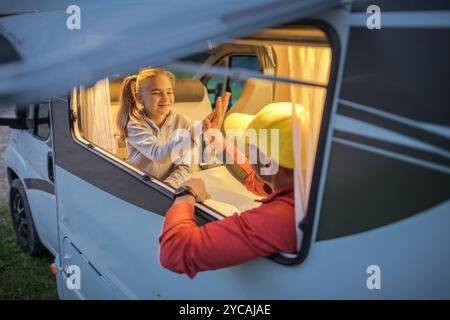 Un père partage un moment joyeux avec sa fille alors qu'ils profitent de moments de qualité ensemble dans un camping-car au crépuscule, entouré par la beauté de la nature. Banque D'Images
