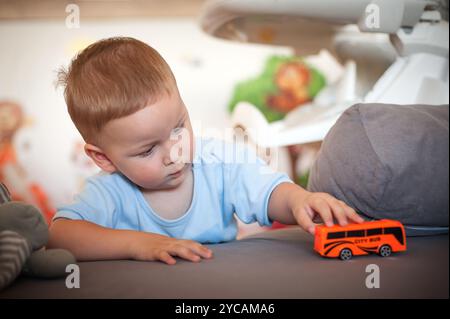 Un tout-petit se concentre intentionnellement sur un petit bus jouet, le déplaçant d'avant en arrière sur une surface douce tout en étant entouré d'un environnement joyeux et coloré designe Banque D'Images