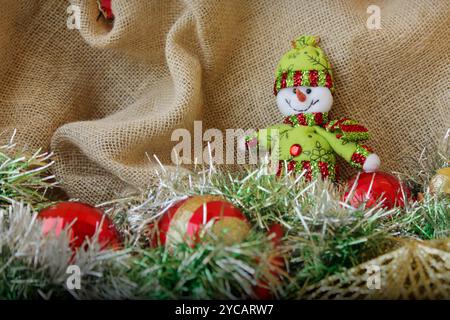 Bonhomme de neige solitaire portant une tenue confortable en laine verte devant un fond de tissu de jute. Boules de Noël rouges et vertes pour la décoration des fêtes de décembre. Banque D'Images