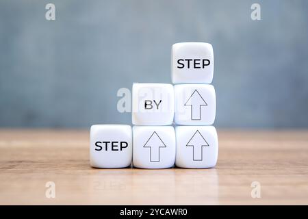 Des blocs blancs portant les mots « STEP BY STEP » et des flèches pointant vers le haut sont empilés sur une table en bois. Banque D'Images