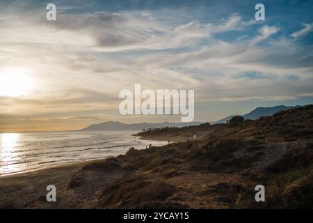 La lumière dorée du coucher de soleil baigne les dunes côtières tandis que les montagnes lointaines offrent une toile de fond magnifique. Le littoral paisible ajoute à la tranquillité et Banque D'Images