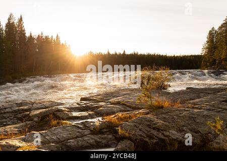 Une scène automnale tranquille en Suède avec un coucher de soleil doré projetant une lumière chaude sur une rivière rapide et un paysage boisé. Banque D'Images