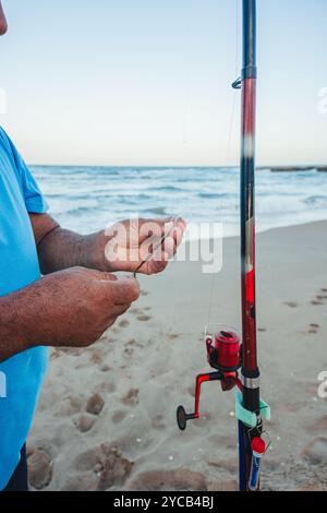 Gros plan d'un homme âgé préparant habilement sa ligne de pêche sur une plage tranquille de Valence au coucher du soleil, incarnant l'esprit intemporel du traditi Banque D'Images